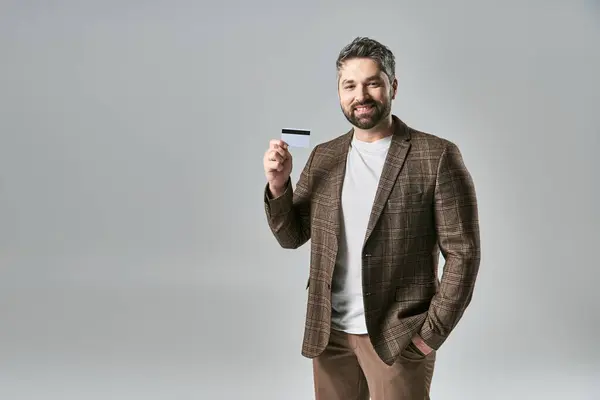 Un homme à la mode avec une barbe frappant une pose tout en tenant une carte dans une tenue élégante sur un fond de studio gris. — Photo de stock