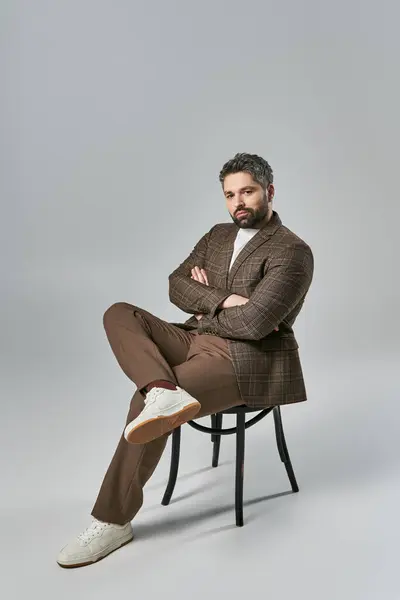 A bearded man sits with arms crossed, exuding strength and poise in elegant attire against a grey studio backdrop. — Stock Photo