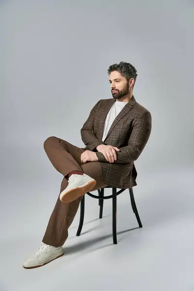 A sophisticated man with a beard sits elegantly on a chair, crossing his legs, in a studio with a grey background. — Stock Photo