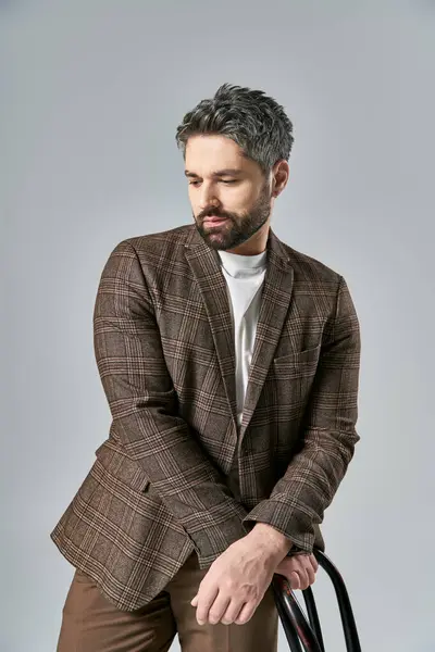 A bearded man in a brown jacket and pants poses with a black chair in a studio setting against a grey background. — Stock Photo