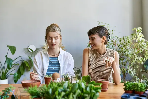Una pareja lesbiana cariñosa, sentada en una mesa en un estudio de arte, rodeada de plantas en maceta. - foto de stock