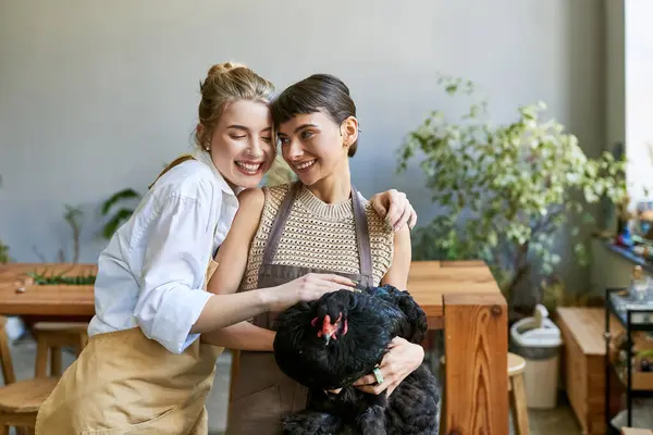 Dos mujeres, una pareja lesbiana cariñosa, de pie en un estudio de arte, sosteniendo la gallina. - foto de stock