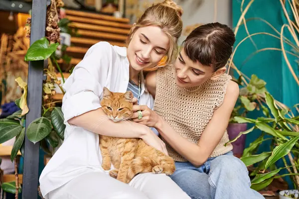 Due donne, una coppia lesbica amorevole, si siedono tranquillamente su un portico con un gatto, circondate da decorazioni artistiche.. — Foto stock