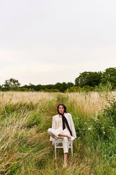 Una bella giovane donna vestita di bianco si siede su una sedia, immersa nella serenità, godendo della brezza estiva in mezzo a un campo panoramico. — Foto stock
