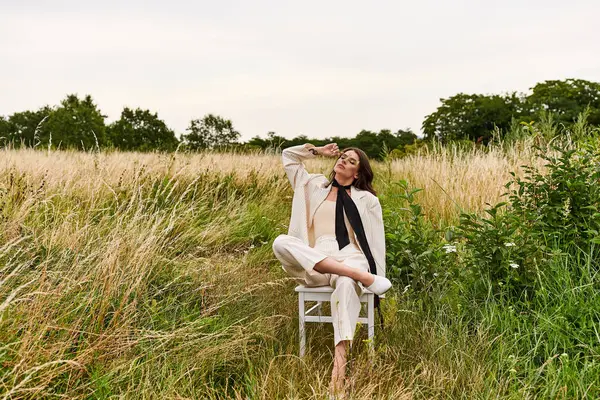 Eine junge Frau, weiß gekleidet, sitzt friedlich auf einem Stuhl inmitten eines üppigen Feldes und tränkt die Wärme der Sommerbrise.. — Stockfoto