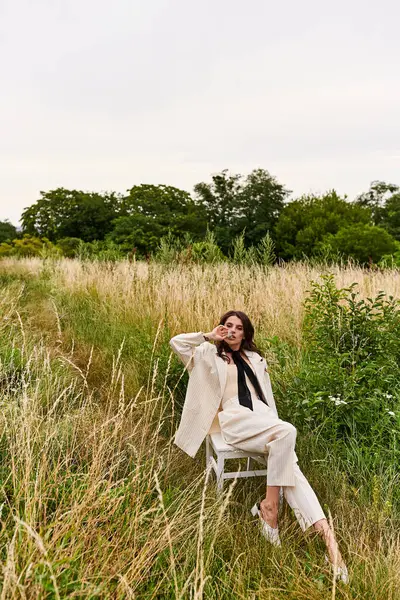 Eine schöne junge Frau in weißem Gewand sitzt auf einem Stuhl in einem sonnenverwöhnten Feld und genießt gelassen die Sommerbrise. — Stockfoto