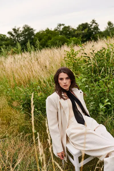 Una hermosa joven vestida de blanco sentada tranquilamente en una silla, empapando la brisa del verano en un exuberante campo. - foto de stock