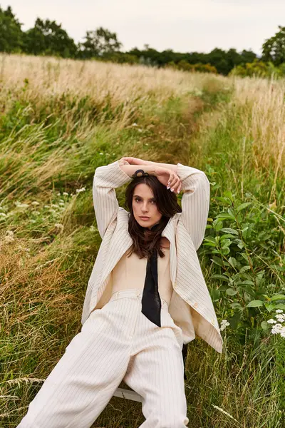 Una hermosa joven vestida de blanco sentada en un campo, con las manos en la cabeza, acariciando la brisa del verano. - foto de stock