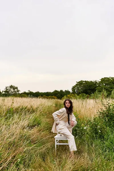 Красивая молодая женщина в белом сидит изящно на стуле, наслаждаясь летним бризом в пышном поле. — стоковое фото