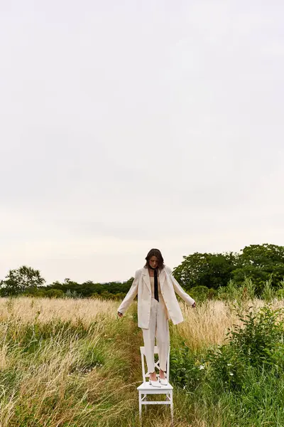 Uma bela jovem mulher de traje branco fica em uma cadeira em um campo, pacificamente encharcada na brisa de verão. Instrução. — Fotografia de Stock