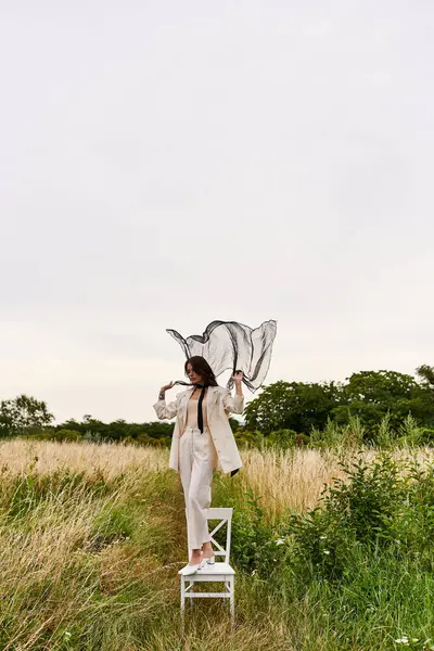 Una elegante joven vestida de blanco se para en una silla, disfrutando de la brisa del verano en un paisaje de campo. - foto de stock