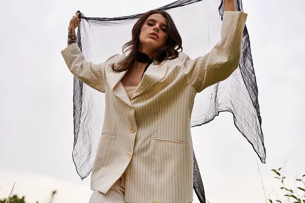 A beautiful young woman in a white suit delicately holds a black shawl while standing in a tranquil field, embracing the summer breeze. - foto de stock
