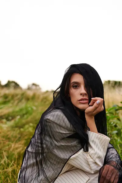 A beautiful young woman in white attire sitting in the grass, thoughtful look with hand on chin, enjoying the summer breeze. - foto de stock