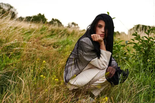 A beautiful young woman in white attire kneels gracefully in a field of tall grass, enjoying the summer breeze. — Stock Photo