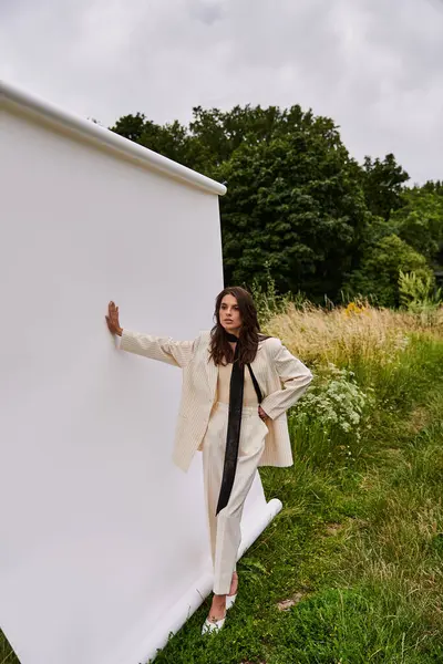 Una joven vestida de blanco se apoya contra una pared blanca, abrazando la brisa del verano en un entorno de campo sereno. - foto de stock