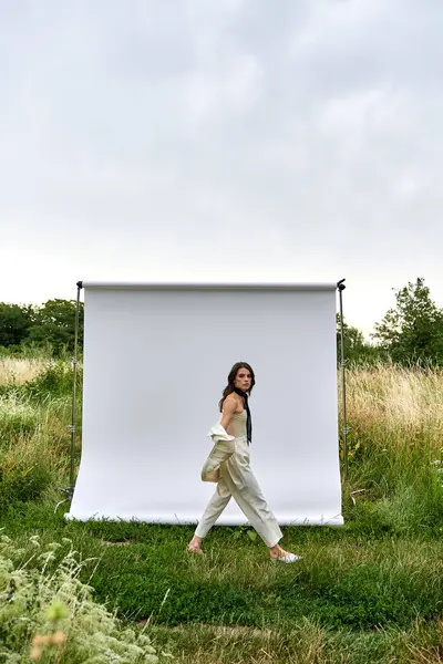 Uma bela jovem mulher de traje branco caminha graciosamente na frente de um pano de fundo branco, apreciando a brisa de verão em um campo. — Fotografia de Stock