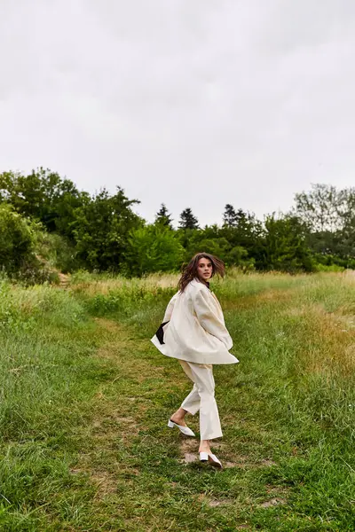 Une belle jeune femme en robe blanche marchant gracieusement à travers un champ luxuriant, se prélassant dans la brise d'été. — Photo de stock