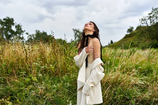 Una hermosa joven vestida de blanco de pie en un campo con los ojos cerrados, empapada en el calor del sol. - foto de stock