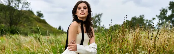 A beautiful young woman in white attire stands serenely in a field of tall grass, embracing the summer breeze. — Photo de stock