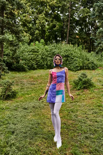 Une belle jeune femme dans une robe colorée et des lunettes de soleil se tient gracieusement dans un champ, profitant de la brise d'été. — Photo de stock