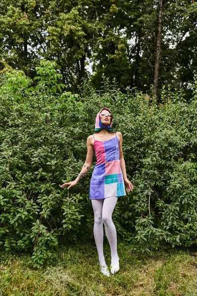 Uma bela jovem mulher em um vestido vibrante e óculos de sol, apreciando a brisa de verão, fica elegantemente na exuberante grama verde. — Fotografia de Stock