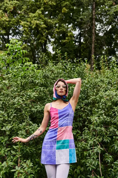 Потрясающая женщина в ярком платье и солнцезащитных очках изящно стоит на живописном поле, наслаждаясь нежным летним бризом. — стоковое фото