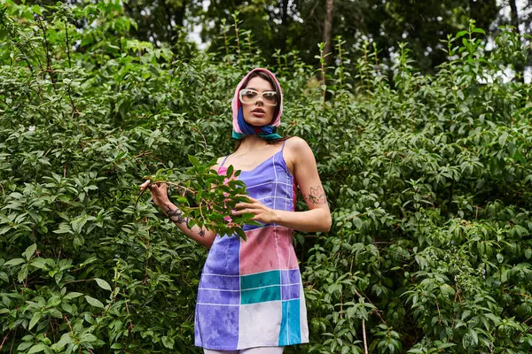 Une jeune femme vibrante dans une robe colorée et des lunettes de soleil se tient devant un buisson, tenant délicatement une plante dans sa main. — Photo de stock