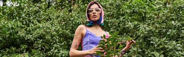 Une jeune femme vibrante dans une robe colorée et des lunettes de soleil se tient devant un buisson, tenant une plante dans sa main, bannière — Photo de stock