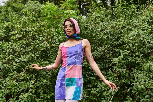 Una hermosa joven se para frente a un arbusto, vistiendo un vestido colorido y gafas de sol, disfrutando de una brisa de verano en la naturaleza. - foto de stock