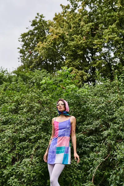 Красивая молодая женщина в ярком платье и солнечных очках наслаждается летним бризом перед пышными деревьями. — стоковое фото