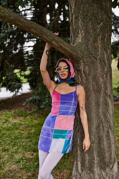 Une belle jeune femme dans une robe vibrante et des lunettes de soleil se tient à côté d'un arbre, profitant de la brise d'été. — Photo de stock