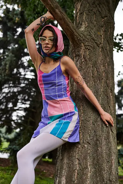 Una hermosa joven con un vestido colorido se apoya contra un árbol, absorbiendo la brisa del verano y la tranquilidad. - foto de stock