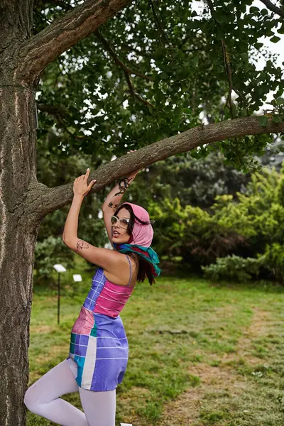 A young woman in a vibrant dress and sunglasses climbing up a tree branch, embracing the summer breeze in nature. - foto de stock