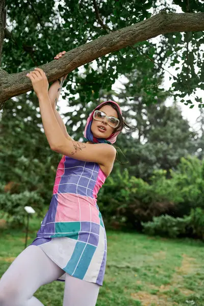 A beautiful young woman in a vibrant dress and sunglasses balancing gracefully on a tree branch, enjoying the summer breeze. — Stock Photo
