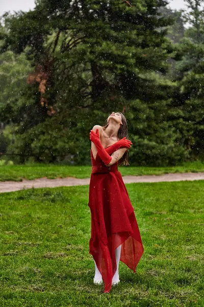 Eine bezaubernde junge Frau in einem leuchtend roten Kleid steht anmutig im Regen und umarmt die Elemente mit Gelassenheit und Eleganz. — Stockfoto