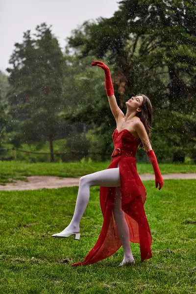 Une jeune femme dans une robe rouge magnifique danse gracieusement sous la pluie, sentant la brise d'été. — Photo de stock