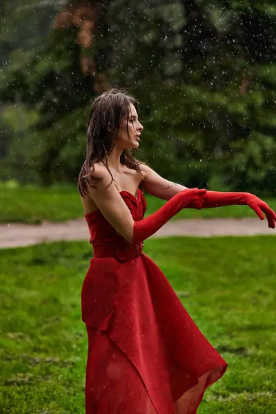 Una joven con un vestido rojo fluido y guantes largos baila con gracia en la lluvia de verano, encarnando aplomo y elegancia. - foto de stock
