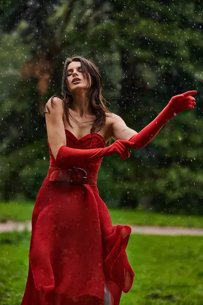 Uma jovem mulher em um vestido vermelho impressionante fica graciosamente em um chuveiro de chuva de verão, abraçando beleza naturezas. — Fotografia de Stock