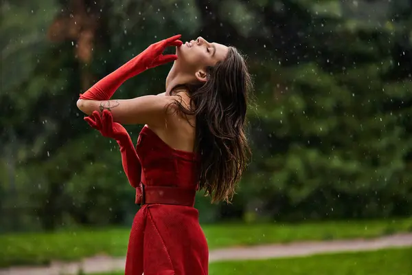Una mujer joven con estilo en un vestido rojo que fluye se levanta con gracia bajo la lluvia que cae, abrazando la serena belleza de la naturaleza. - foto de stock