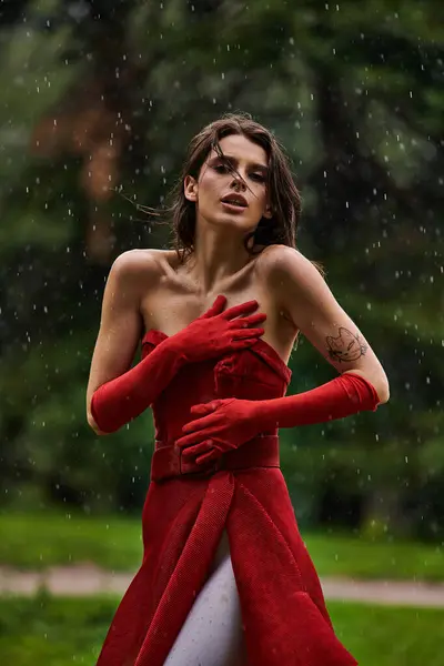 A stunning young woman in a red dress and gloves stands gracefully in the rain, embracing the elements with poise and beauty. — Stock Photo