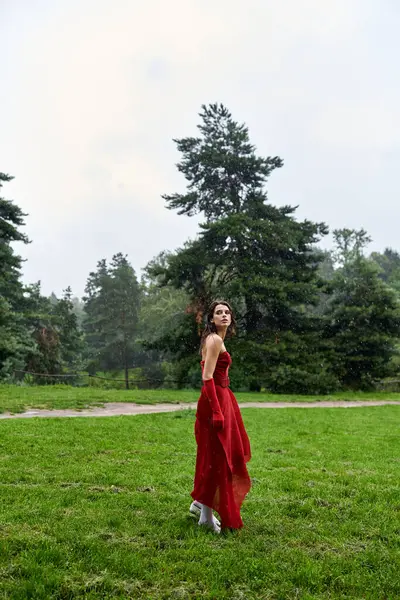 An attractive young woman in a striking red dress and long gloves gracefully stands in a sunlit field, feeling the summer breeze. — Stock Photo