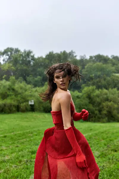An elegant young woman in a flowing red dress and long gloves stands gracefully in a sunlit field, soaking in the summer breeze. - foto de stock