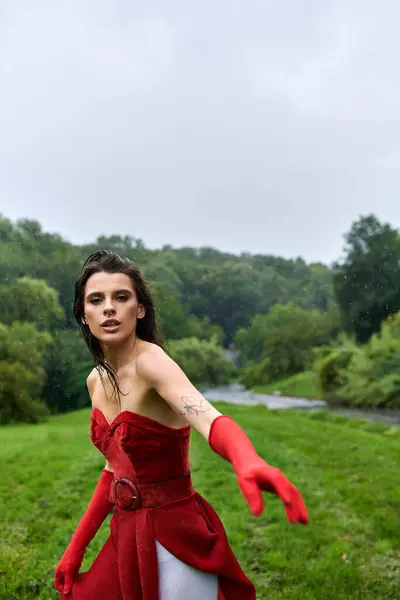 An attractive young woman in a flowing red dress and long gloves stands gracefully in a lush green field, enjoying the summer breeze. — Stock Photo