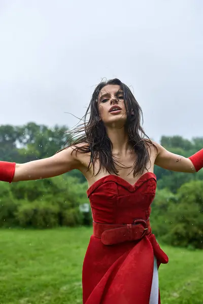 A stunning young woman in a red dress and long gloves stands gracefully in a field, feeling the gentle summer breeze. — Stock Photo