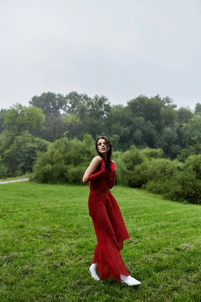 Una giovane donna elegante in un abito rosso vibrante e guanti lunghi in piedi con grazia in un campo sereno, accarezzando la calda brezza estiva. — Foto stock