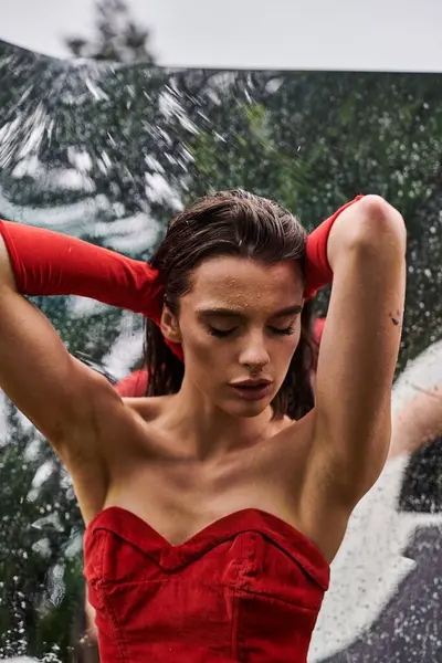 Una mujer con un llamativo vestido rojo y guantes largos se levanta elegantemente bajo la lluvia, disfrutando del aguacero del verano.. - foto de stock