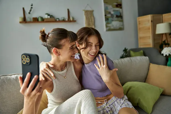 Two women, a beautiful lesbian couple, sitting on a couch joyfully taking a selfie together. — Stock Photo