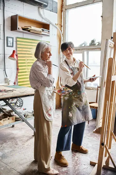 A mature lesbian couple standing beside an easel in an art studio. — Stock Photo