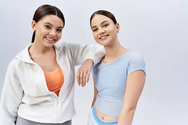 Dos guapas adolescentes morenas vestidas con ropa deportiva se paran una al lado de la otra en una pose simétrica contra un fondo gris de estudio. - foto de stock