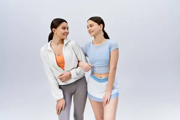 Дві красиві брюнетки молоді жінки в спортивному вбранні стоять поруч, виключаючи впевненість і дружбу на сірому студійному фоні. — стокове фото